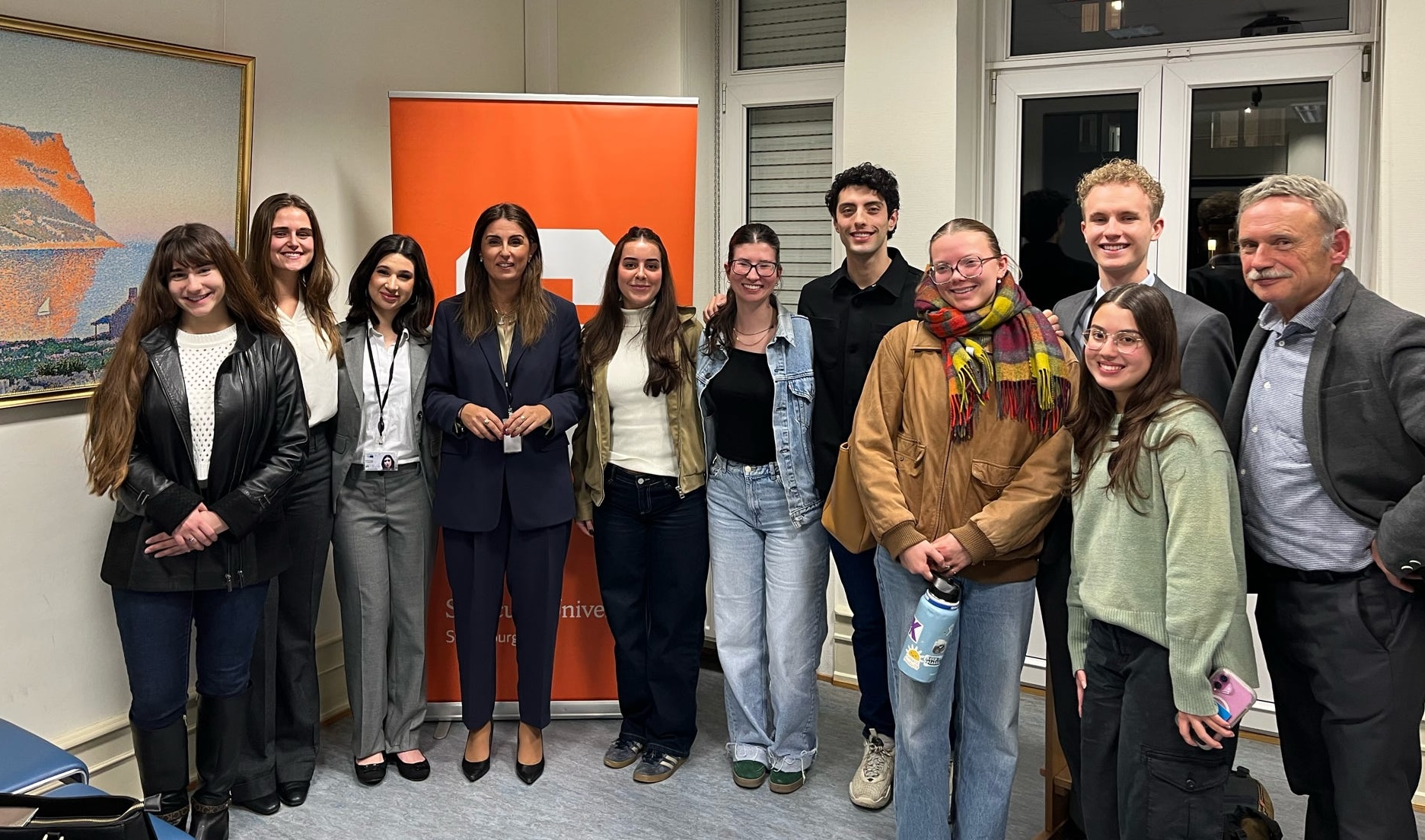 A group of people standing together in a room in front of an orange banner, with chairs and artwork visible.