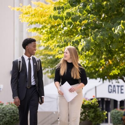 Two students dressed in business attire walk and talk on a university campus, holding papers near a tree-lined walkway.