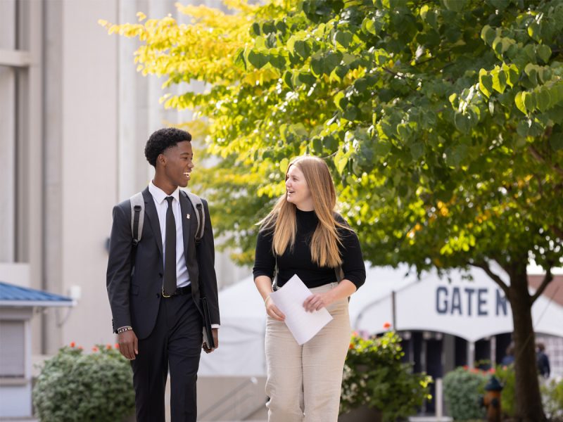 Two students dressed in business attire walk and talk on a university campus, holding papers near a tree-lined walkway.