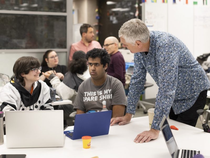 A group of people work together in a classroom-style space, with one person leaning over a table to assist others who are using laptops.