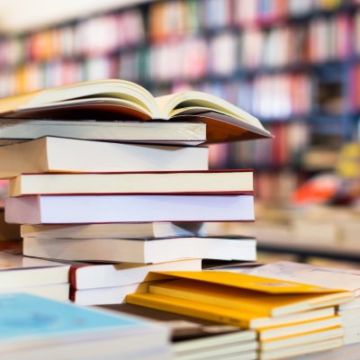 Stacks of books on a table with an open book on top, in a bookstore filled with shelves of colorful books