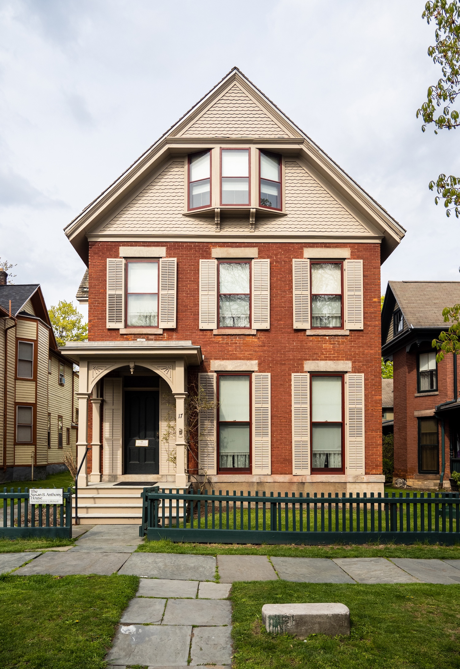 A tall, three‑story brick house with light‑colored shutters, a covered front porch, and a green picket fence, viewed from the sidewalk.