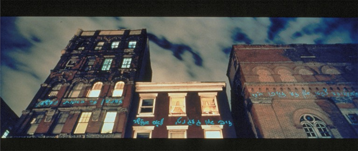 Yiddish text projected in blue laser light across the facades of brick tenement buildings on Manhattan's Lower East Side at dusk, in Shimon Attie's "Lasers Writing Out (in Yiddish) Jewish Senior's Sleeping Dream"