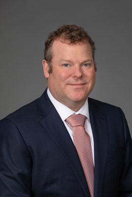 Headshot of man wearing a navy suit with an orange patterned tie against a gray background.