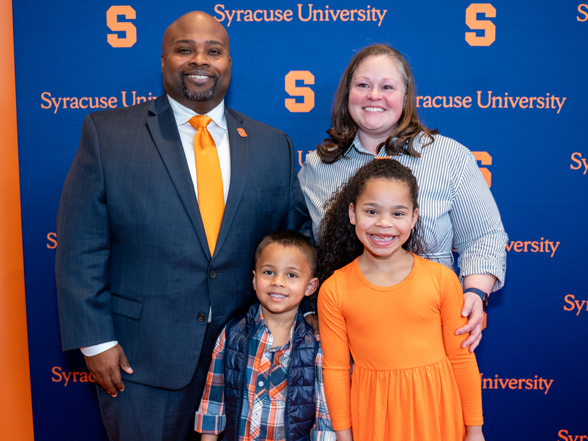 A person poses with their wife and their two children in front of a Syracuse University step-and-repeat banner.
