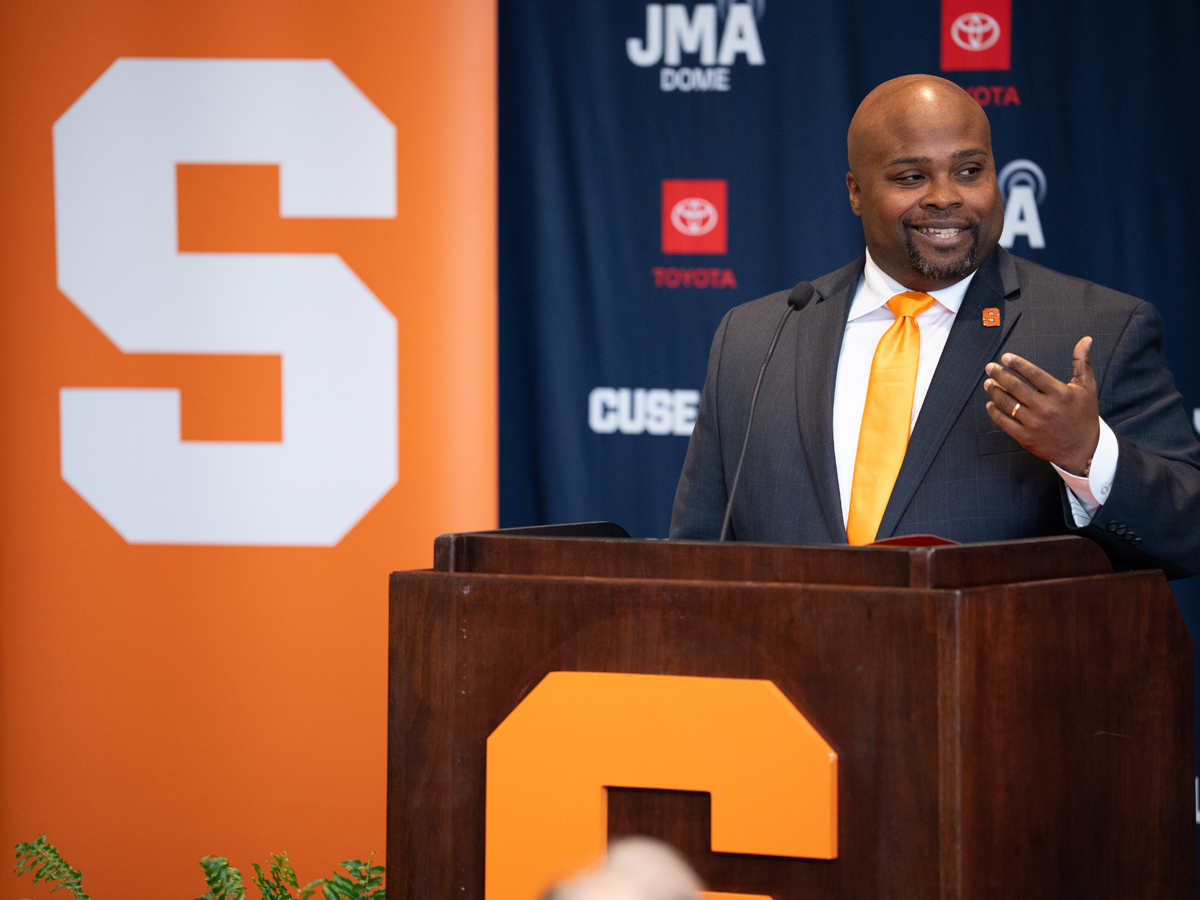 A person speaks at a podium during an introductory press conference as Syracuse University athletics director, with the block S logo visible behind him.