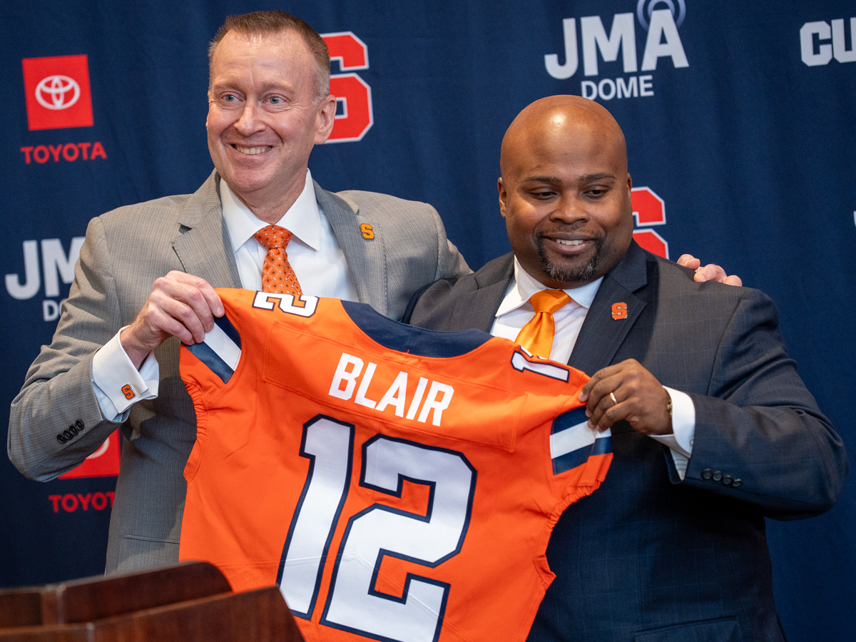 Two people stand holding an Orange number 12 Syracuse football jersey with the name Blair in white lettering on the back.