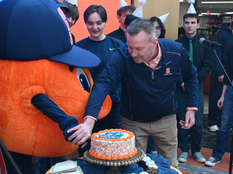 A person cuts an orange-and-blue decorated cake beside a large orange mascot during a campus celebration, with attendees in the background.