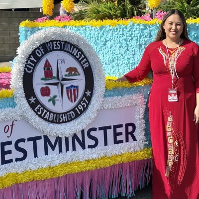 A person in a red dress standing beside the City of Westminster parade float.