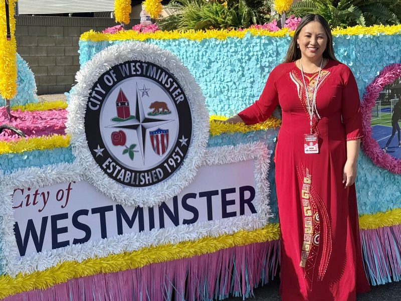 A person in a red dress standing beside the City of Westminster parade float.