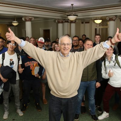 A smiling person stands at the front of a group of diverse students. They are in a casual setting that appears to be indoors. Some students are wearing clothing with various university logos.