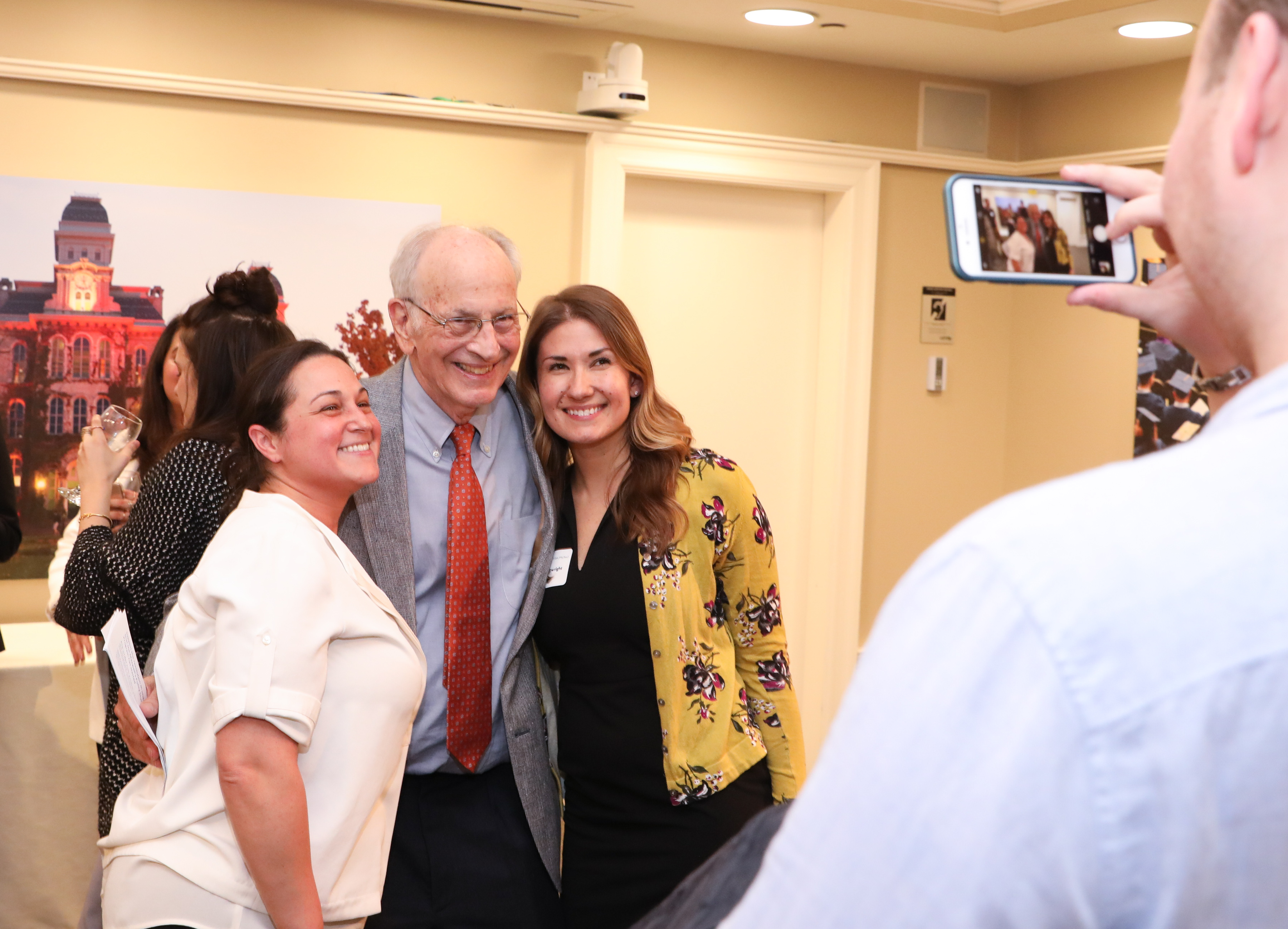 A group of people smiling for a photo, with one person taking the picture on a smartphone. They are indoors, and in the background, there is a picture of a building.
