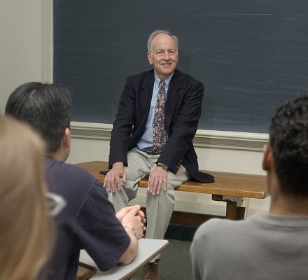 A person sits on a desk at the front of a classroom, smiling and engaged in conversation with students seated in front of him. A chalkboard is visible in the background.