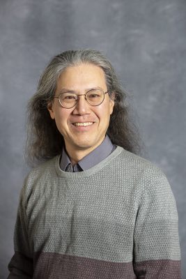 Headshot of person wearing glasses and a gray two-tone sweater over a collared shirt against a gray background.
