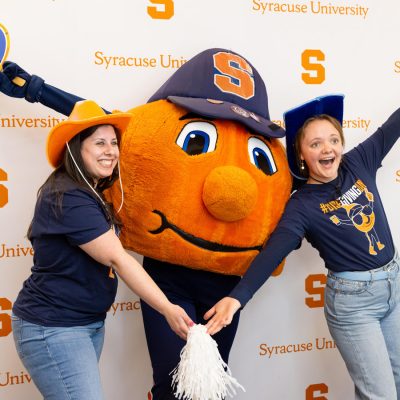 Two smiling women in Syracuse University navy blue t-shirts and jeans pose enthusiastically with Otto the Orange, Syracuse University's mascot — a large orange character wearing a navy cap with an "S" logo. One woman wears an orange cowboy hat and holds up an Otto fan sign; the other raises her arm excitedly. They stand in front of a Syracuse University branded step-and-repeat backdrop with orange "S" logos.
