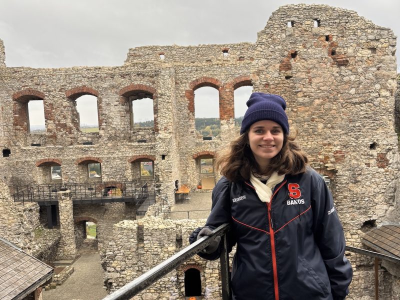 Person wearing a dark jacket and knit hat standing on a metal railing inside the ruins of a large stone fortress with multiple arched window openings.