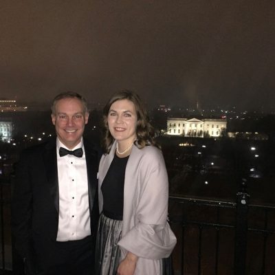 A man in a tuxedo with bow tie and a woman in a black dress with a light pink/mauve wrap and pearl necklace pose together on an outdoor terrace at night, with the illuminated White House visible in the background against the Washington, D.C. skyline.
