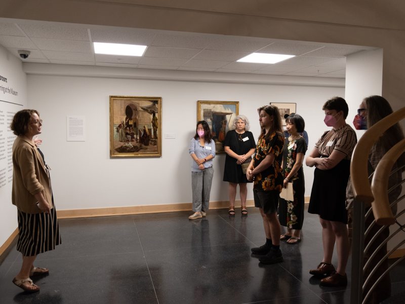 A museum educator addresses a small group of visitors standing before framed paintings in a gallery.