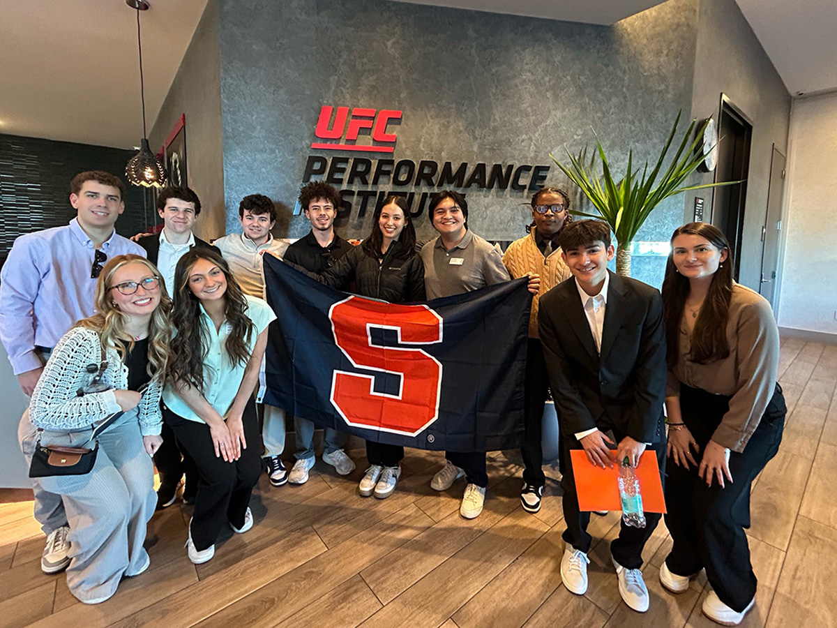 A group of approximately 11 Syracuse University students pose with a Syracuse 'S' flag in the lobby of the UFC Performance Institute.