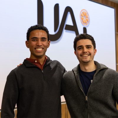 Two men smiling with arms around each other in front of a United AI logo display.
