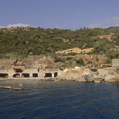 Ruins of a former coastal mining site with arched stone structures along a rocky Mediterranean shoreline.