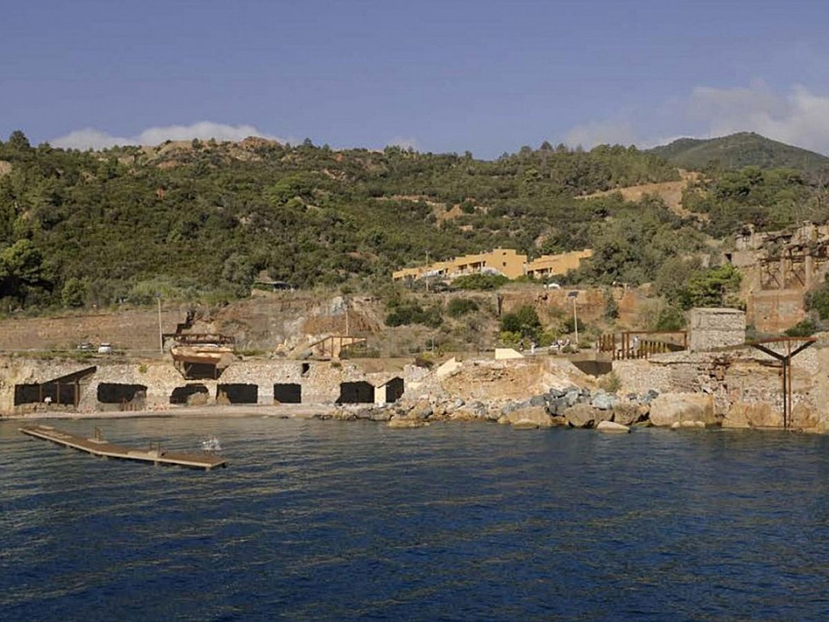 Ruins of a former coastal mining site with arched stone structures along a rocky Mediterranean shoreline.