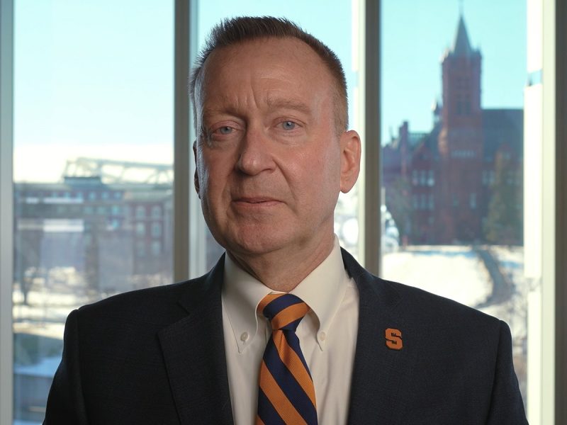 A person in a suit jacket with a Syracuse University "S" lapel pin and striped tie stands before a window overlooking a snow-covered campus, with a red brick building with a tower visible in the background