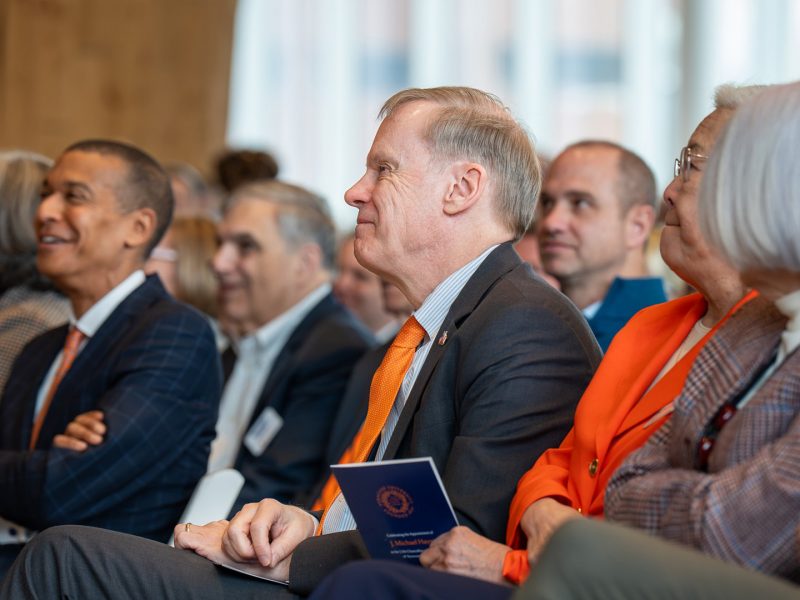 Attendees seated in a row at an indoor press conference, holding event programs and listening to the speaker.