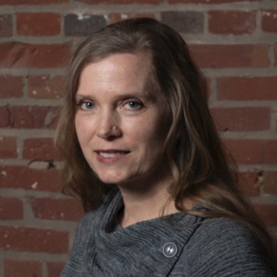 A person with long wavy hair wearing a textured gray top stands in front of a red brick wall.