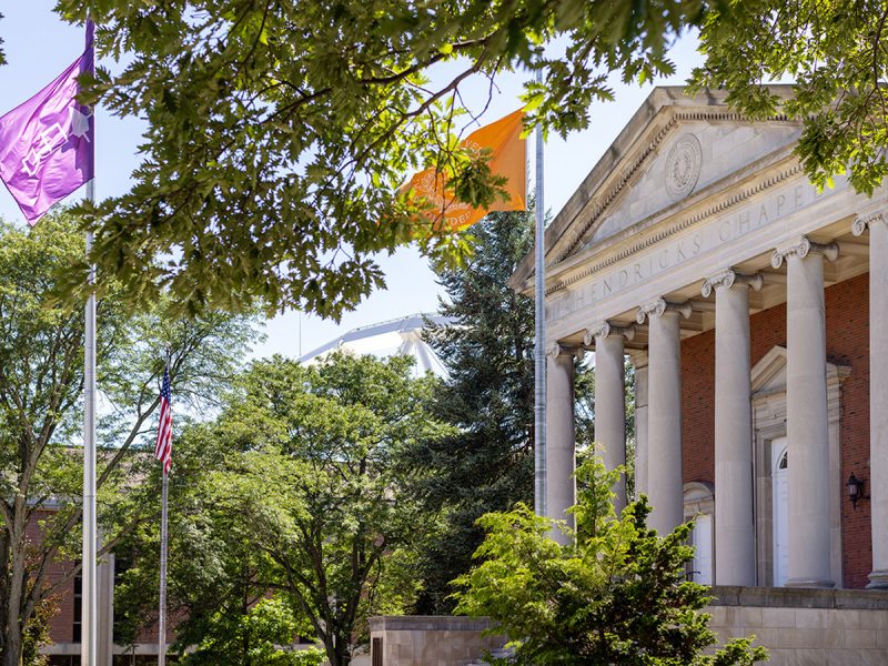 Exterior of Hendricks Chapel surrounded by greenery, Purple Haudosaunee flag and orange University flag