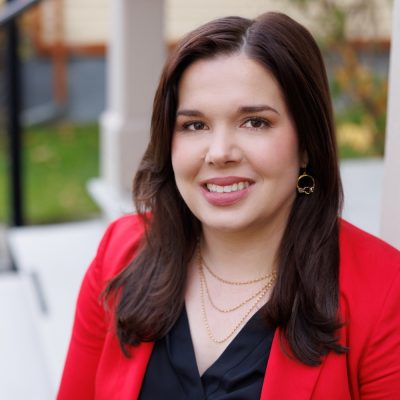 A person with long dark hair, wearing a red blazer and layered gold necklaces, is seated on outdoor steps.