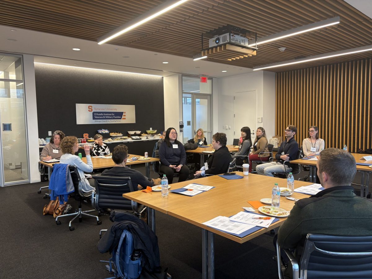 Approximately 15 people are seated at rectangular tables arranged in a U-shape during a workshop session at the D'Aniello Institute for Veterans and Military Families at Syracuse University. A woman at the center of the group is leading a discussion.
