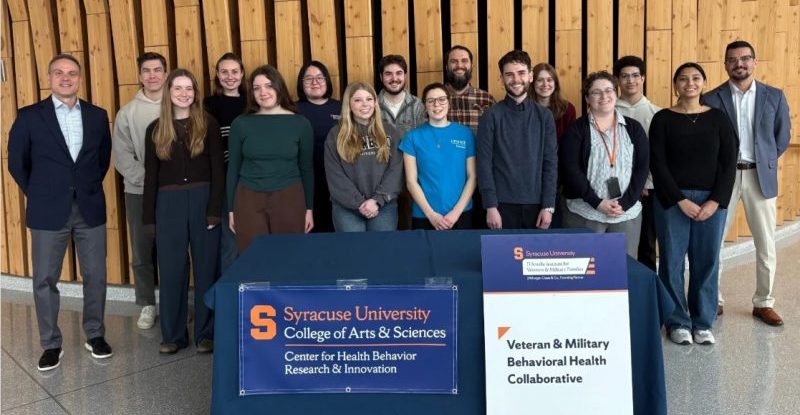 A group of approximately 15 people stand together in front of a wood-paneled wall. In the foreground are two table signs. The sign on the left reads "Syracuse University College of Arts & Sciences, Center for Health Behavior Research & Innovation." The sign on the right, bearing the D'Aniello Institute for Veterans and Military Families logo, reads "Veteran & Military Behavioral Health Collaborative." The group includes faculty, staff and students.