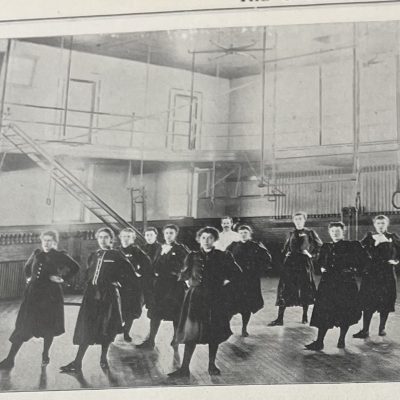 A vintage black‑and‑white photo of a group of people standing in uniform rows inside a large gymnasium with climbing apparatus and railings in the background.