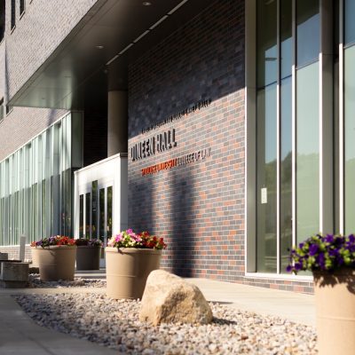 Modern academic building entrance with brick and glass facade, large windows, and planter-lined walkway.