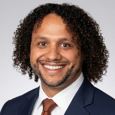 Man in blue jacket, white shirt and orange tie, smiling.