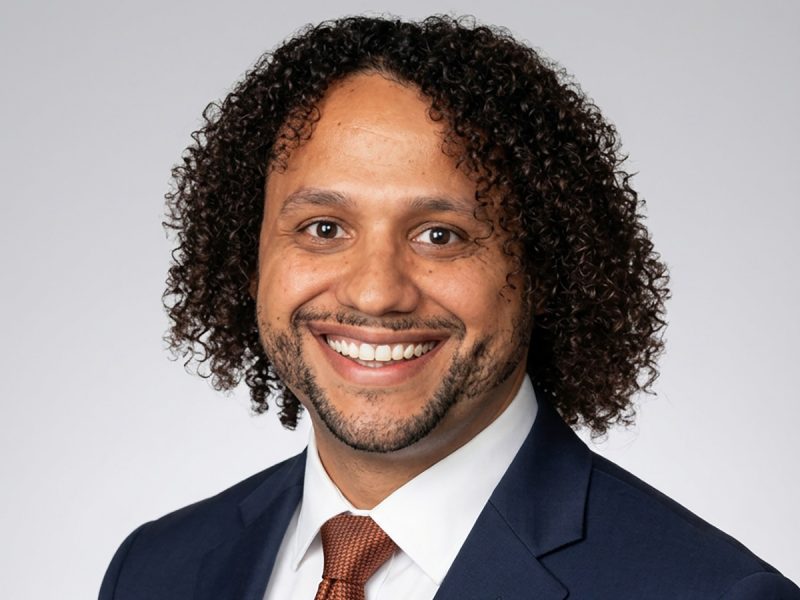 Man in blue jacket, white shirt and orange tie, smiling.