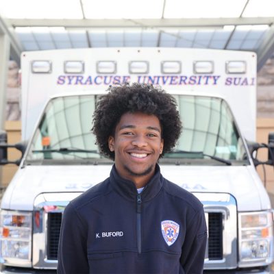 Person in a Syracuse University ambulance service uniform standing in front of a parked Syracuse University SUA1 ambulance