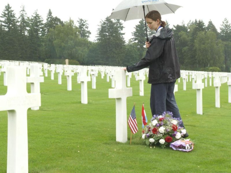 A woman holding an umbrella stands at a grave marker amid rows of white crosses at a military cemetery.