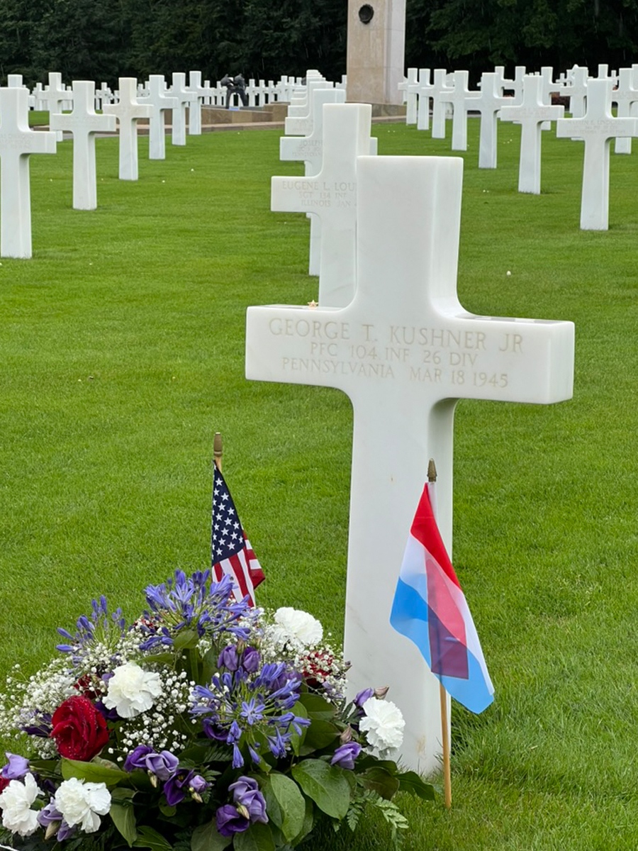 White cross grave marker for George T. Kushner Jr., PFC, 26th Division, adorned with flowers and U.S. and Luxembourg flags.