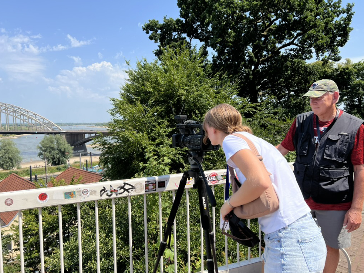 A young woman looks through a video camera on a tripod outdoors overlooking a river and arched bridge.