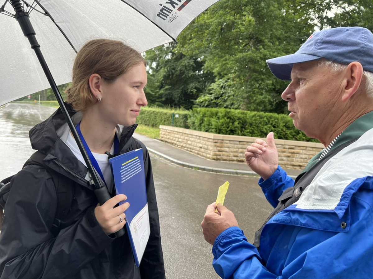 A young woman holding an umbrella and blue folder listens intently to an older man in a rain jacket.