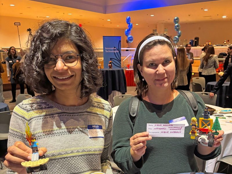 Two women in a brightly room, holding small Lego models they made. Both are smiling.