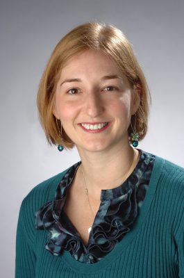Headshot of woman wearing a teal sweater with a ruffled collar and beaded earrings against a gray background.