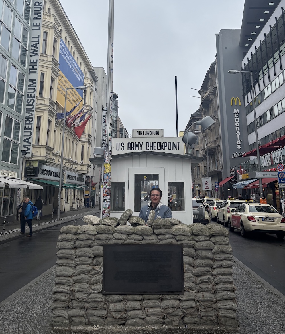 Historic street scene at Checkpoint Charlie with a small U.S. Army guard booth, sandbags in front, and tall buildings lining the road on both sides.