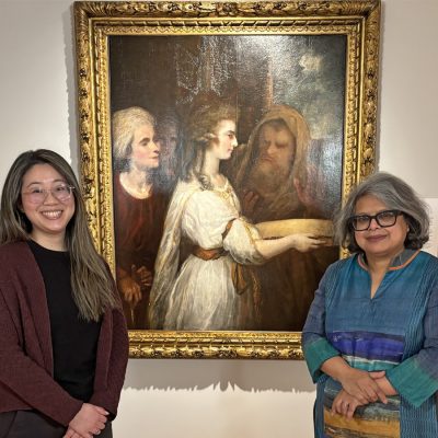 Two people standing in front of a large, framed classical painting displayed on a gallery wall