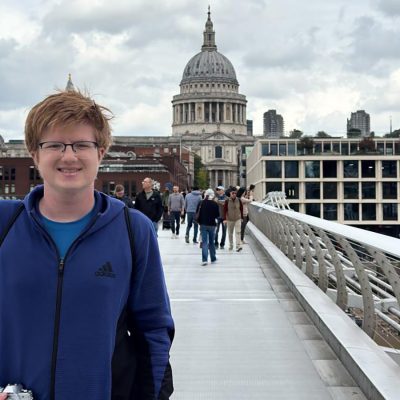 A smiling young person with red hair and glasses stands on London's Millennium Bridge, with St. Paul's Cathedral visible in the background.