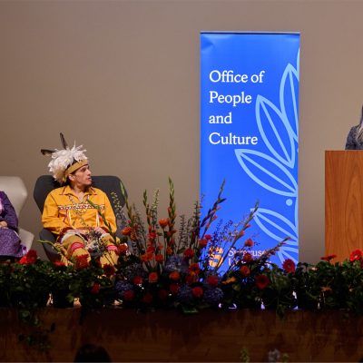 Stage presentation with seated participants and a speaker at a podium beside an ‘Office of People and Culture’ banner