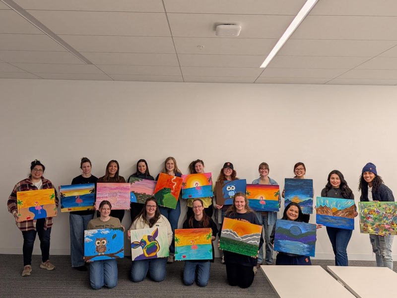 A group of students smile and hold up colorful paintings at a paint and sip event.
