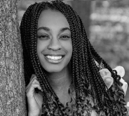A black-and-white portrait of a young woman with long box braids, smiling warmly while leaning against a tree trunk in an outdoor setting.